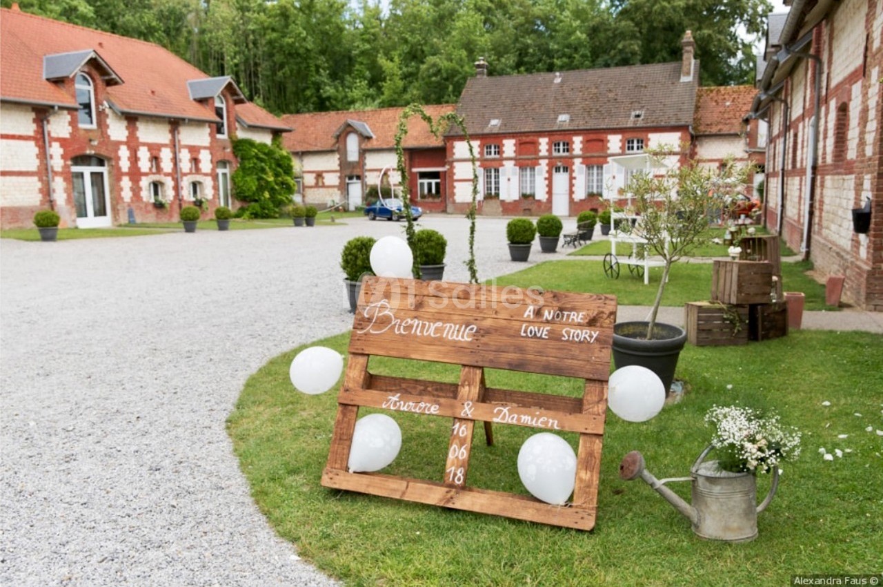 Panneau en bois décoratif avec ballons blancs, placé dans une cour devant des bâtiments en briques et un jardin fleuri.