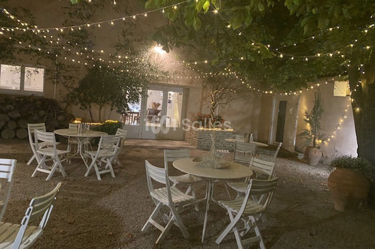 Cour extérieure éclairée par des guirlandes lumineuses, avec des tables et chaises blanches disposées sur un sol gravillonné.