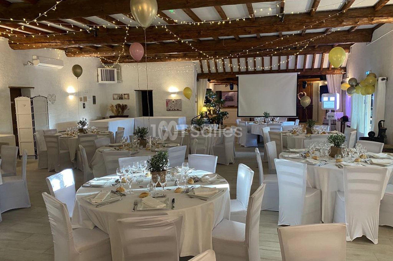 Salle de réception décorée avec des tables rondes dressées, des chaises blanches et des guirlandes lumineuses suspendues.