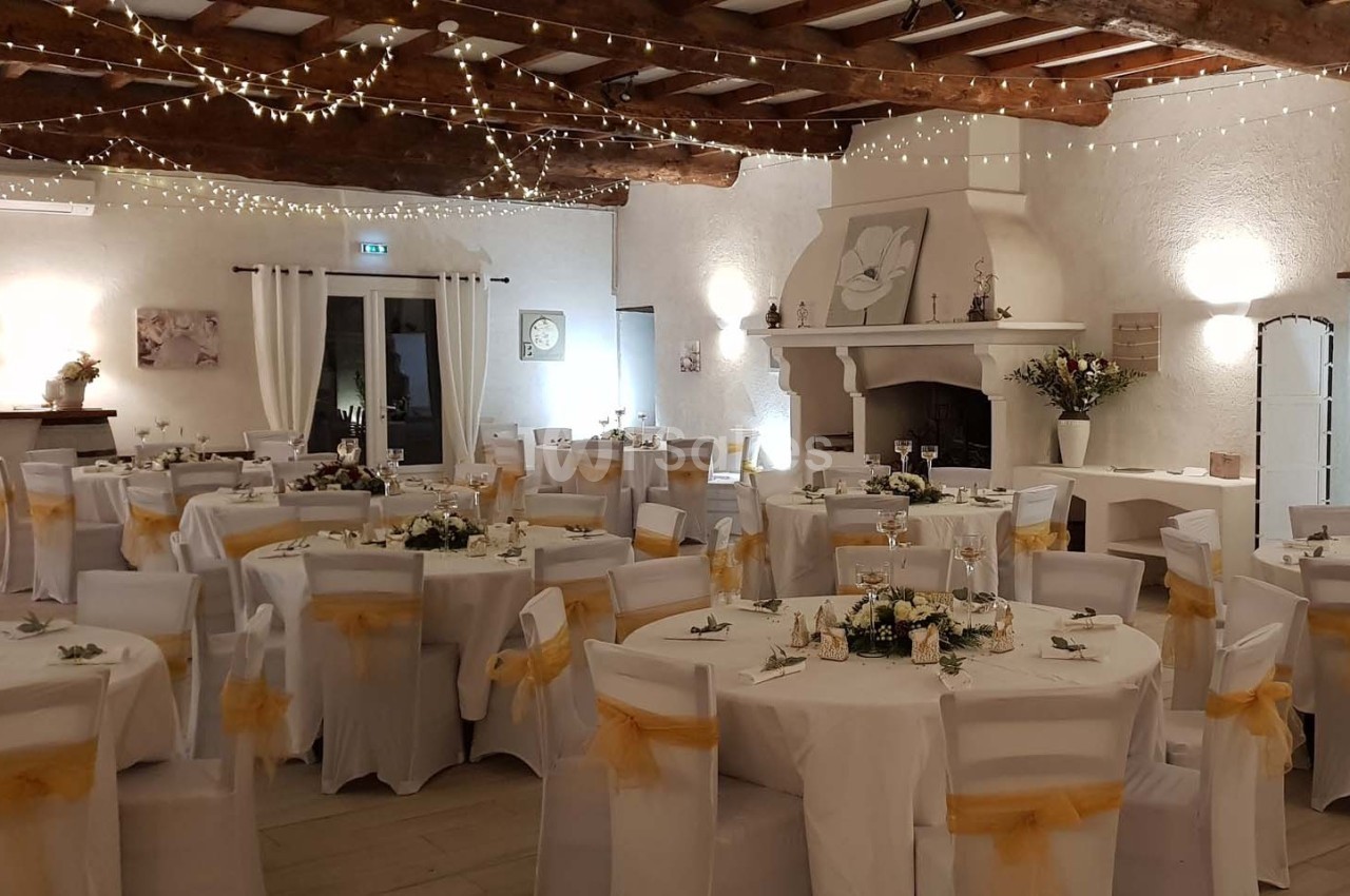 Salle de réception décorée avec des tables rondes, nappes blanches, chaises ornées de nœuds jaunes et guirlandes lumineuses.