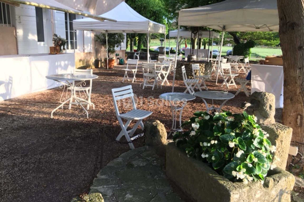 Terrasse extérieure avec tables et chaises blanches sous des tonnelles, entourée de végétation et de fleurs.
