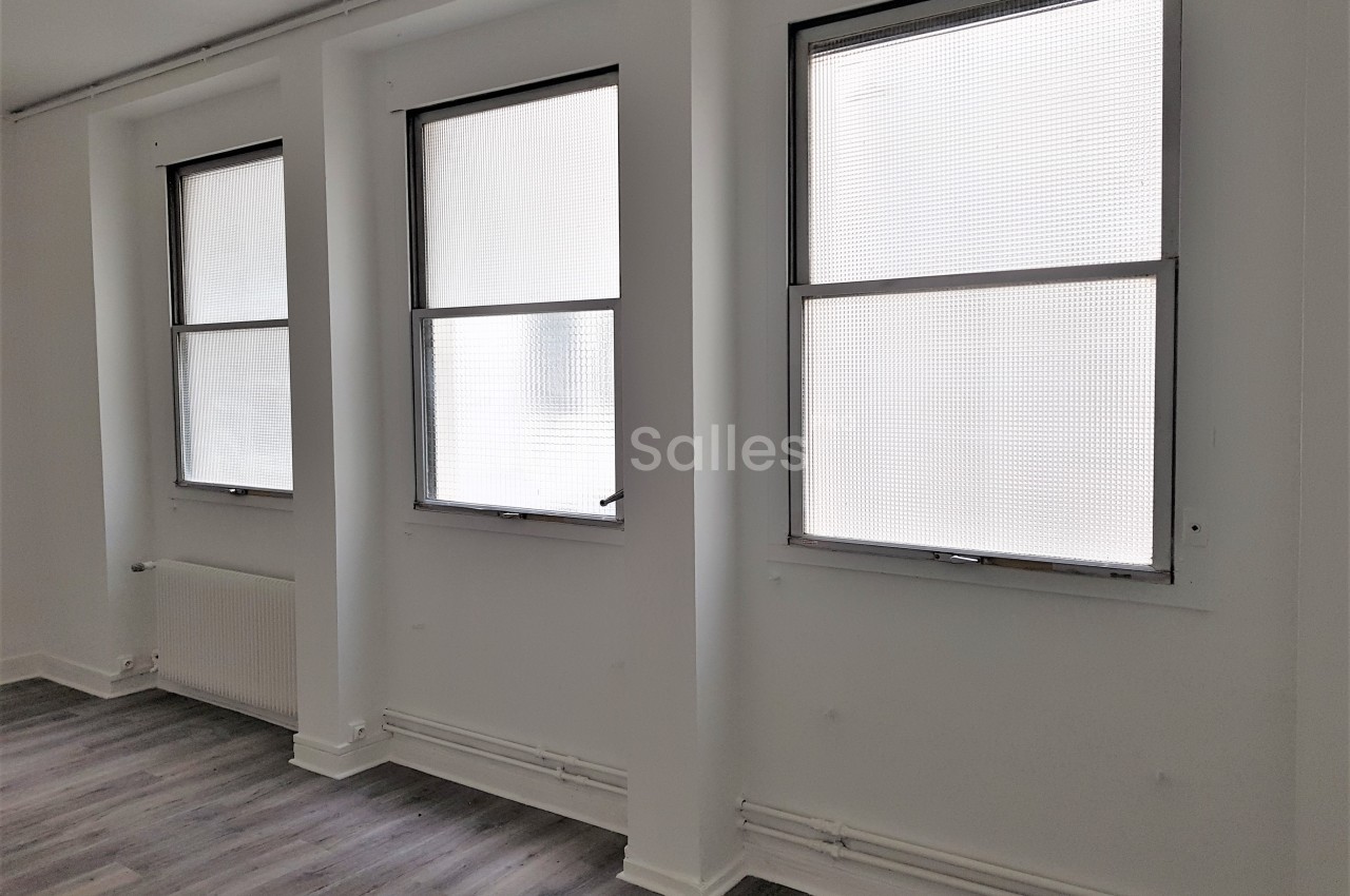 Trois fenêtres alignées dans une pièce lumineuse aux murs blancs et au sol en parquet gris.
