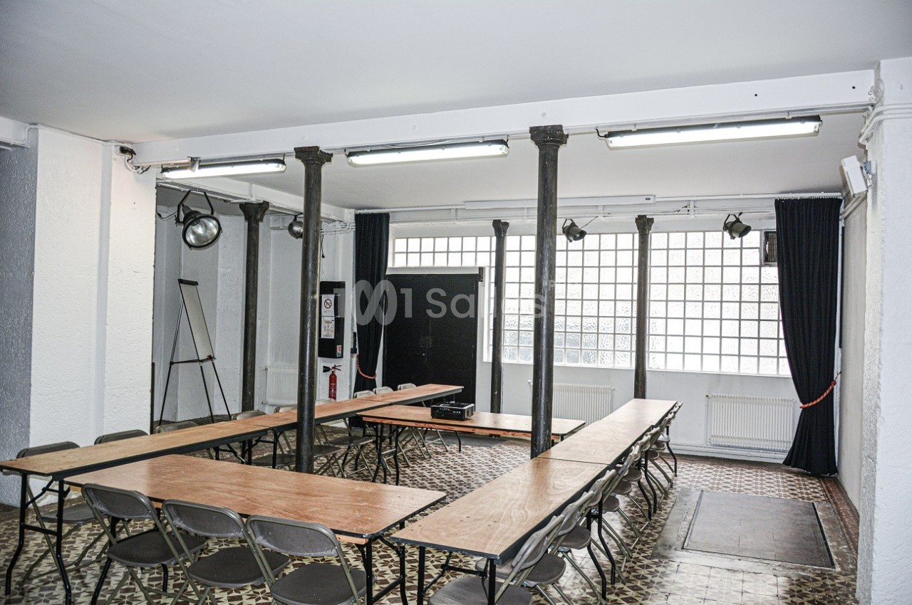 Salle lumineuse avec tables en bois disposées en U, chaises pliantes, colonnes métalliques et grandes fenêtres vitrées.