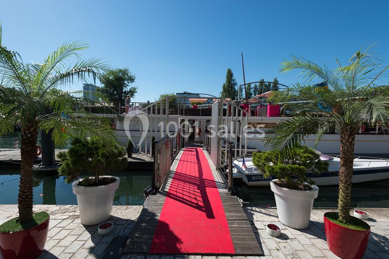 Entrée d'une péniche avec un tapis rouge, entourée de palmiers en pots, amarrée sur un canal par temps ensoleillé.