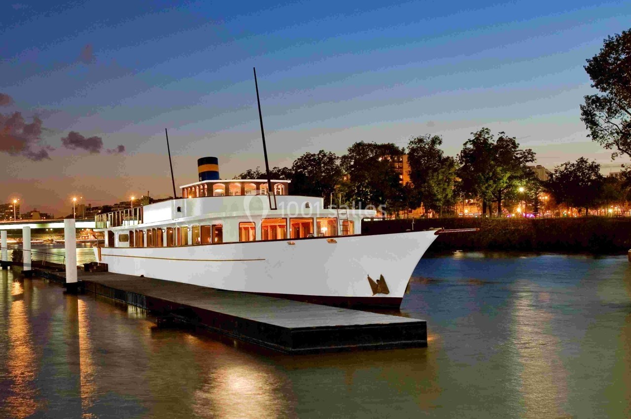 Bateau blanc amarré à un quai sur une rivière au crépuscule, avec des lumières et des arbres en arrière-plan.