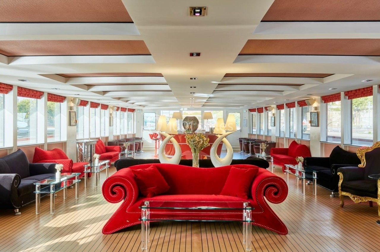 Salon spacieux avec canapé rouge central, fauteuils noirs, tables en verre et grandes fenêtres laissant entrer la lumière.