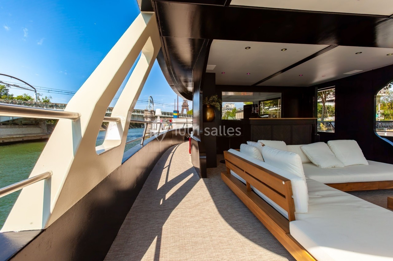 Pont extérieur d'un bateau avec canapé et coussins blancs, offrant une vue sur un canal urbain par temps ensoleillé.