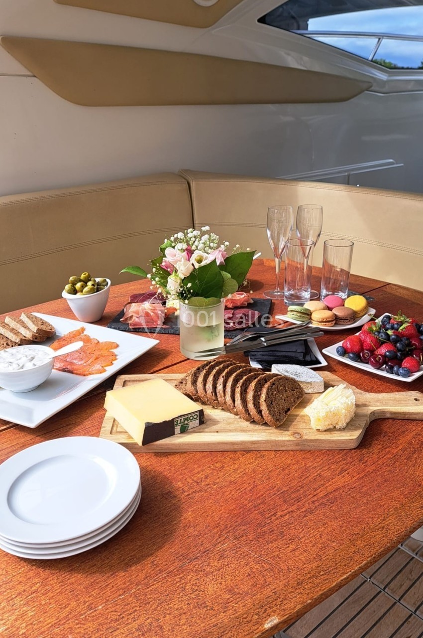 Table en bois avec un repas composé de pain, fromage, saumon, fruits, olives, fleurs et verres à champagne.
