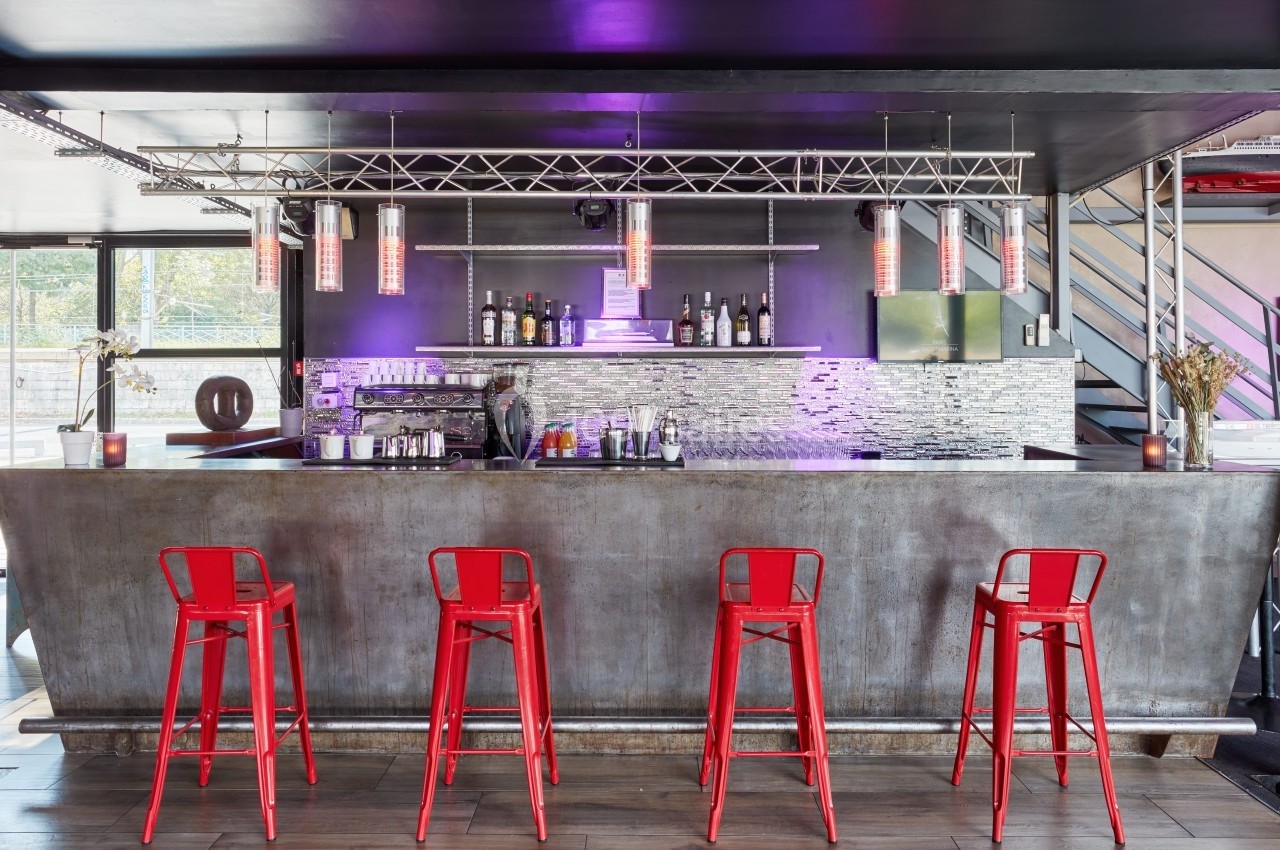 L'Atelier du France Bar moderne avec comptoir métallique, tabourets rouges, étagères de bouteilles et éclairage violet.