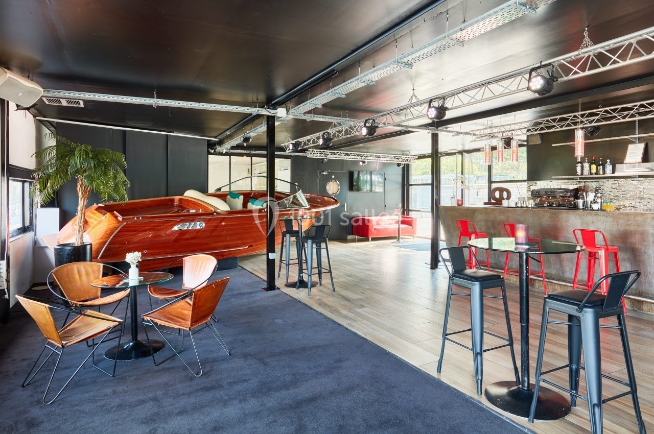 L'Atelier du France Salle moderne avec bar, tables hautes, fauteuils design et un bateau en bois aménagé en élément décoratif.
