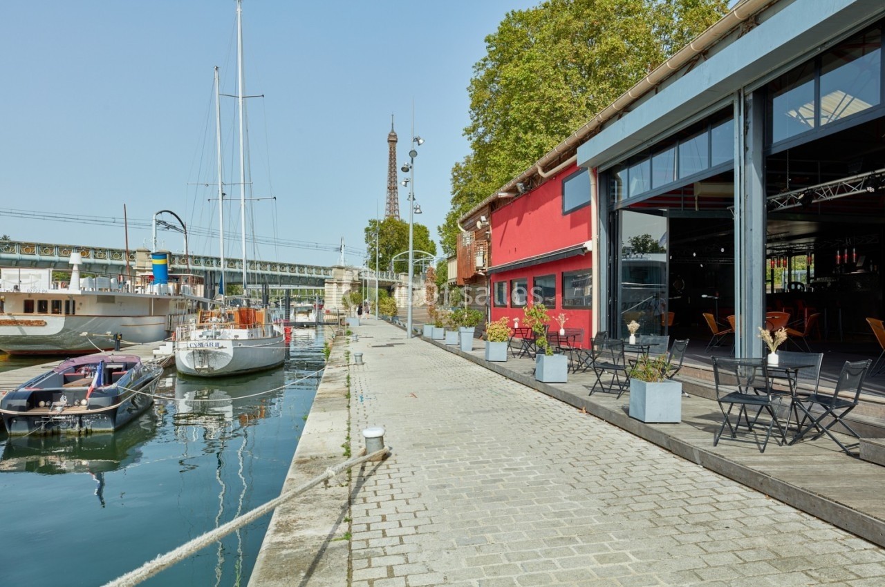 L'Atelier du France Terrasse avec tables et chaises le long d'un quai, près de bateaux amarrés et d'un bâtiment rouge, sous un ciel dégagé.
