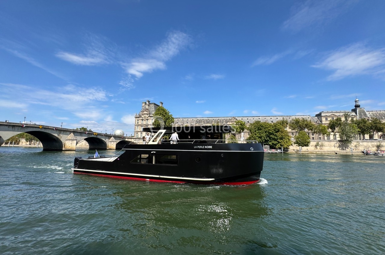 La Perle Noire Bateau noir naviguant sur une rivière, avec un pont et des bâtiments historiques en arrière-plan sous un ciel bleu.