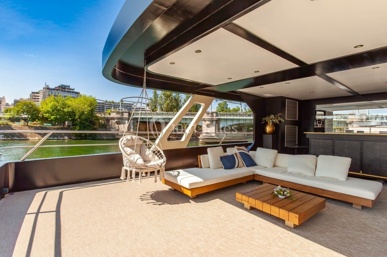 La Perle Noire Terrasse d'un yacht avec canapé blanc, table basse en bois, fauteuil suspendu et vue sur un pont et des bâtiments.