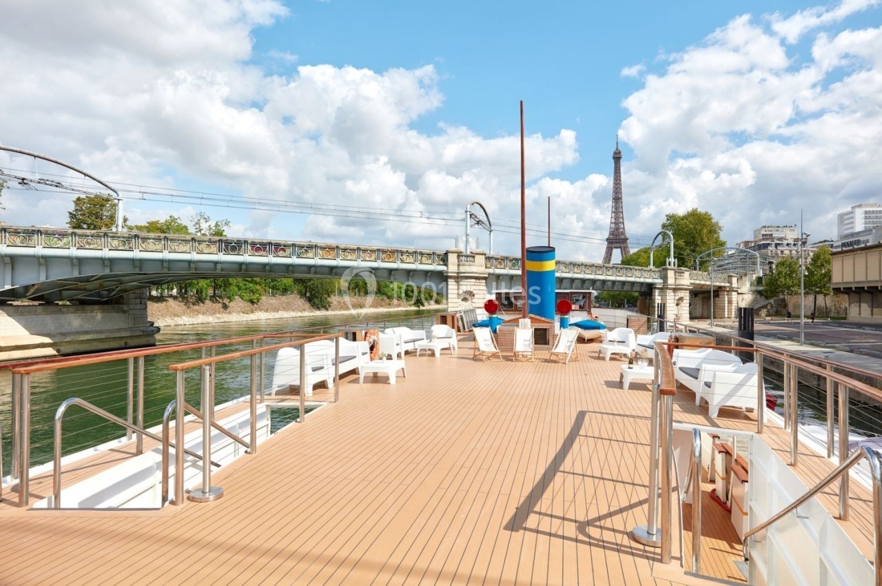 Le Clipper Paris Pont supérieur d'un bateau avec des chaises longues, vue sur la Seine, un pont ferroviaire et la tour Eiffel en arrière-plan.