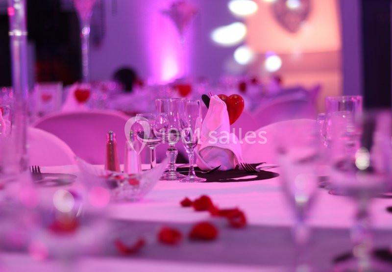 Tables décorées avec nappes blanches, verres, serviettes pliées et pétales rouges dans une ambiance lumineuse rose.