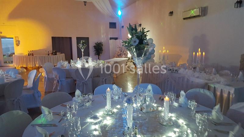 Salle de réception décorée avec des tables rondes, nappes blanches, chandeliers et centre de table floral illuminé.