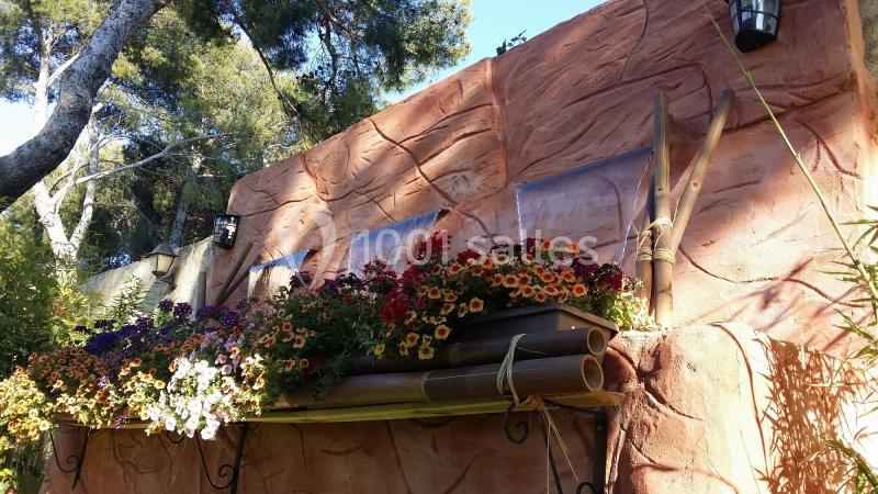 Mur en pierre ocre avec jardinières en bois garnies de fleurs colorées, entouré de végétation et éclairé par la lumière du…