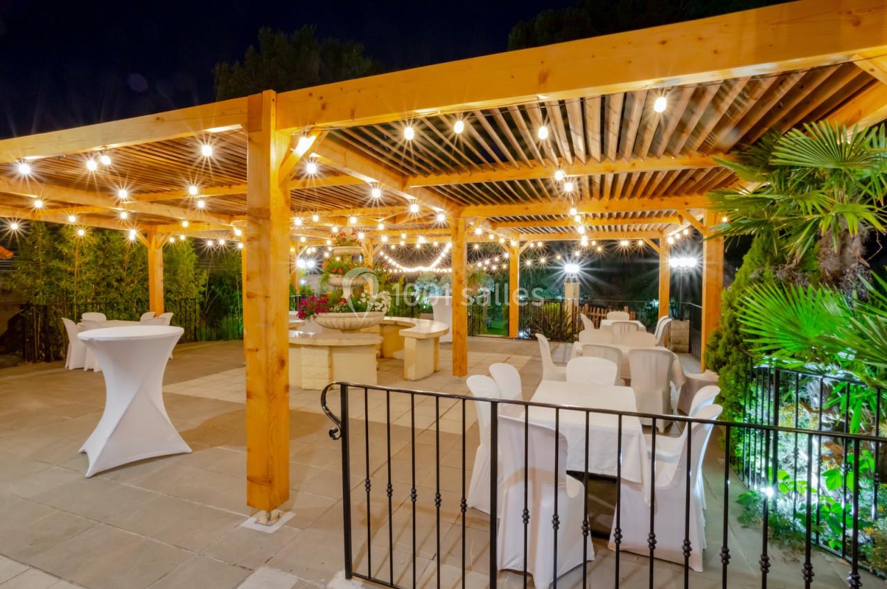 Pergola en bois éclairée par des guirlandes lumineuses, avec tables et chaises blanches dans un espace extérieur de nuit.