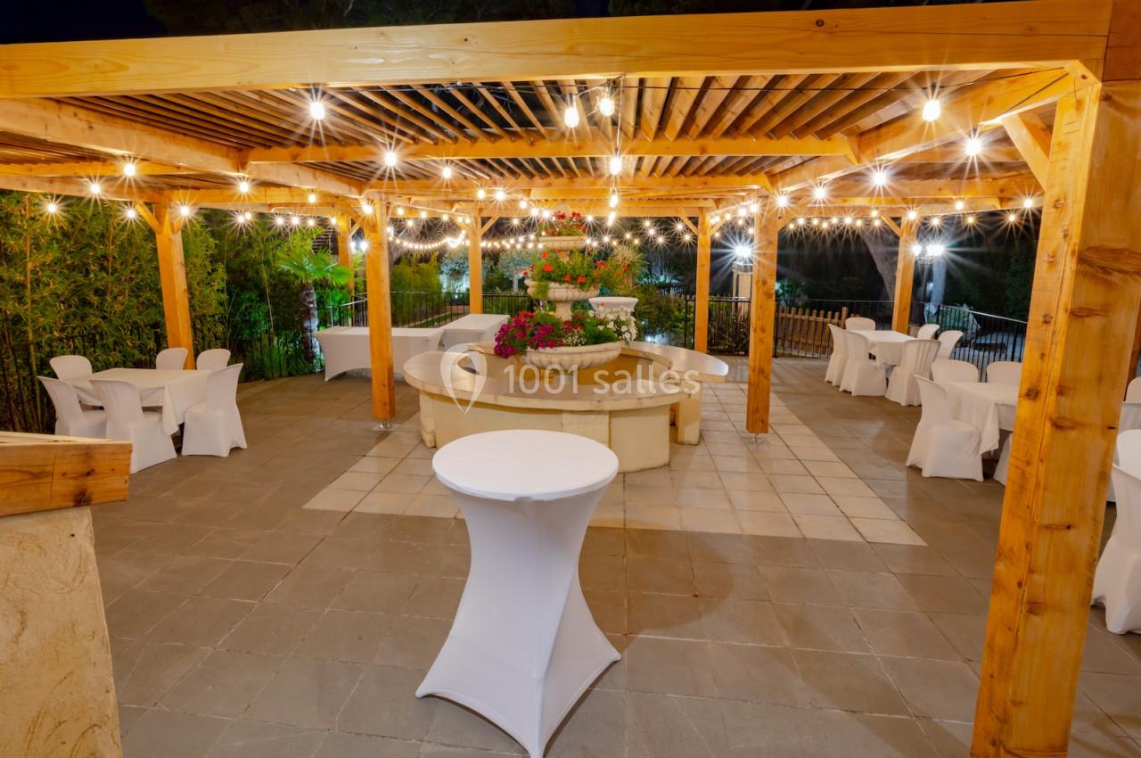 Pergola en bois éclairée par des guirlandes lumineuses, avec des tables et des chaises blanches disposées autour d'une…
