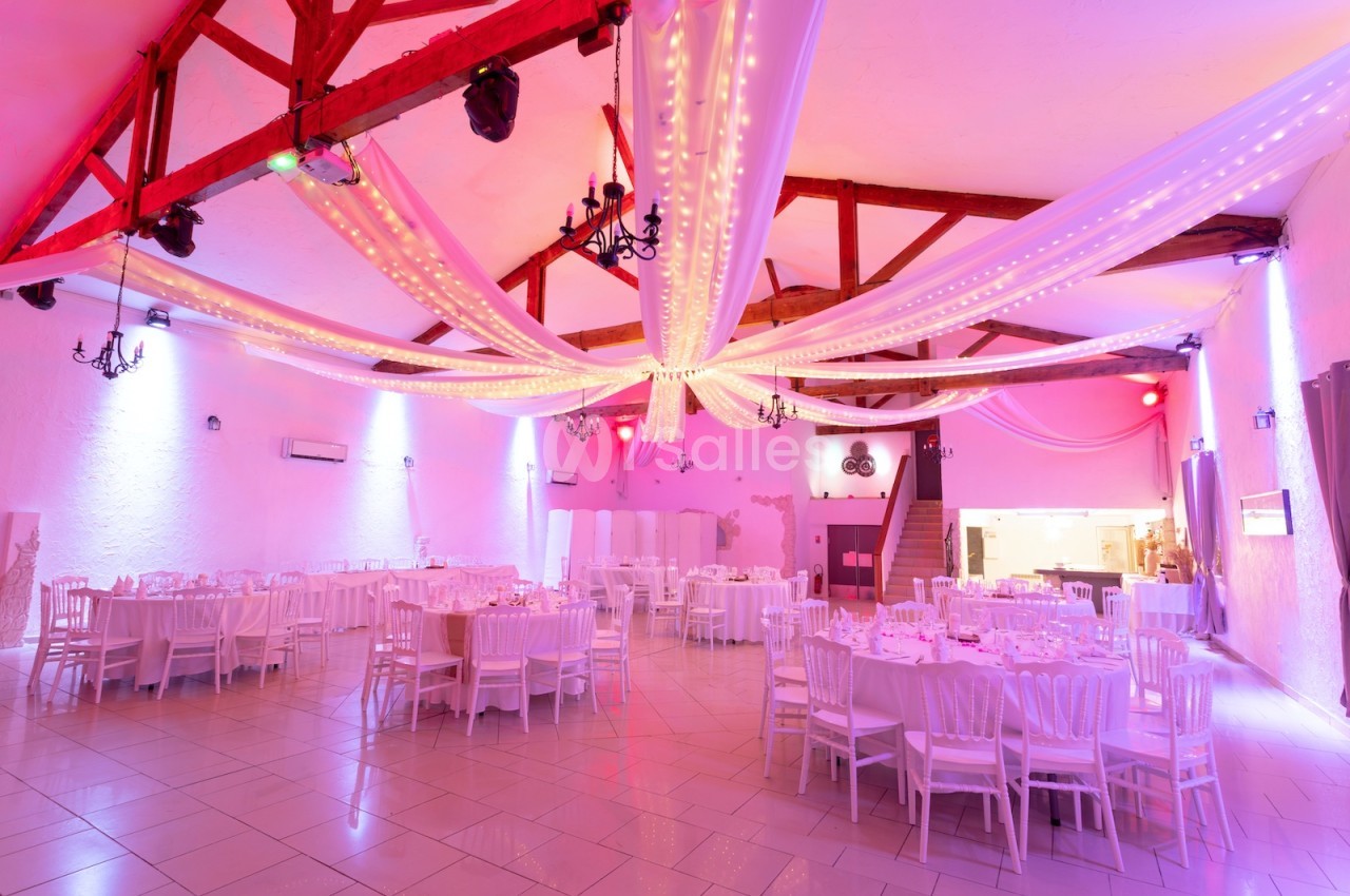 Salle de réception décorée avec des guirlandes lumineuses suspendues, tables rondes dressées et chaises blanches.