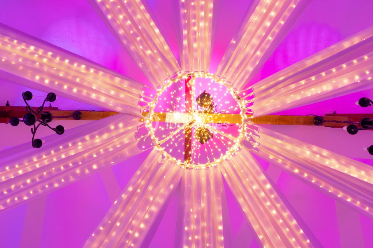 Structure lumineuse circulaire suspendue, ornée de guirlandes LED et de drapés blancs, éclairée en rose.