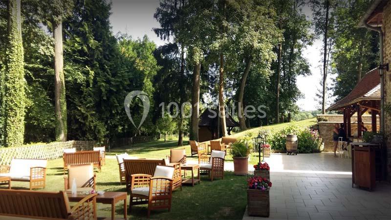 Espace extérieur avec fauteuils en bois, terrasse pavée et vue sur un jardin arboré par une journée ensoleillée.