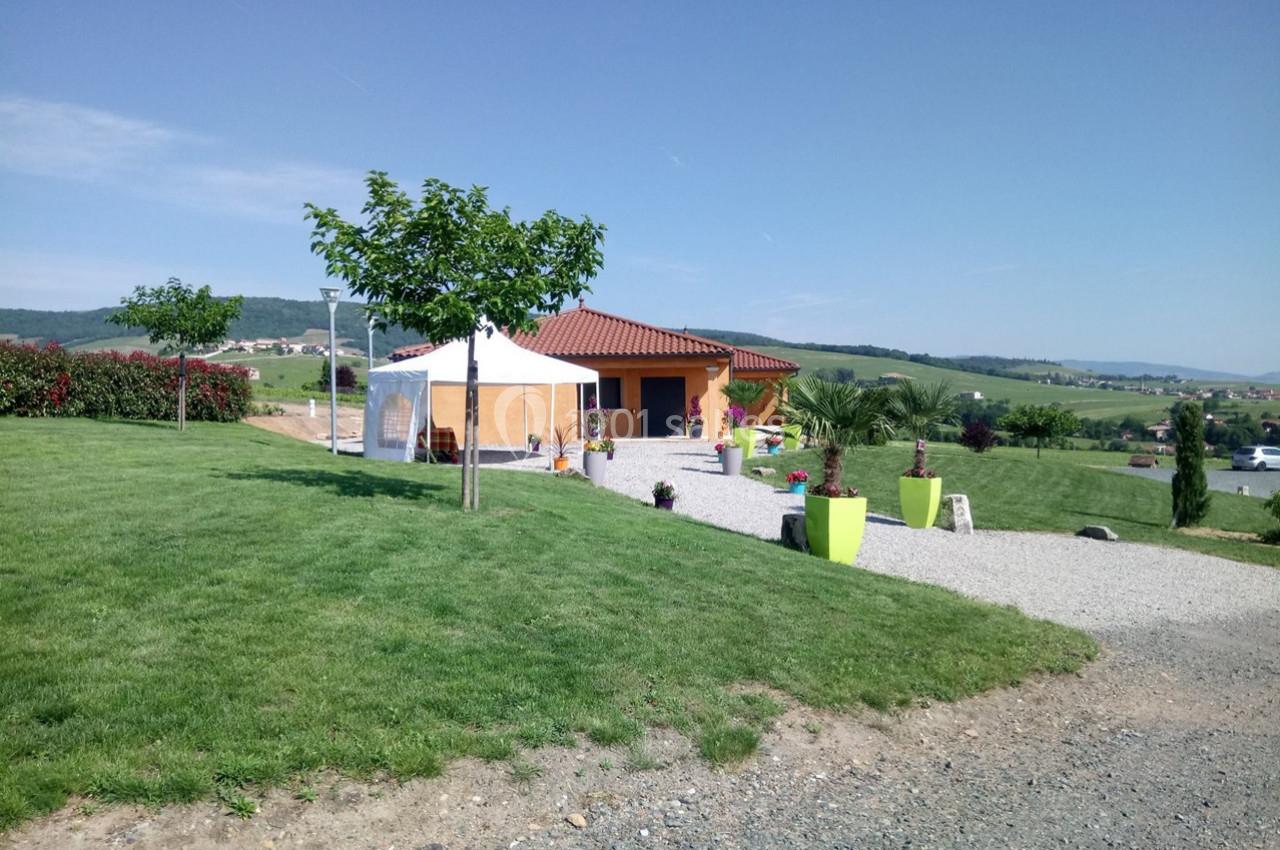 Maison avec toit en tuiles, terrasse, tente blanche et jardin verdoyant dans un paysage rural ensoleillé.