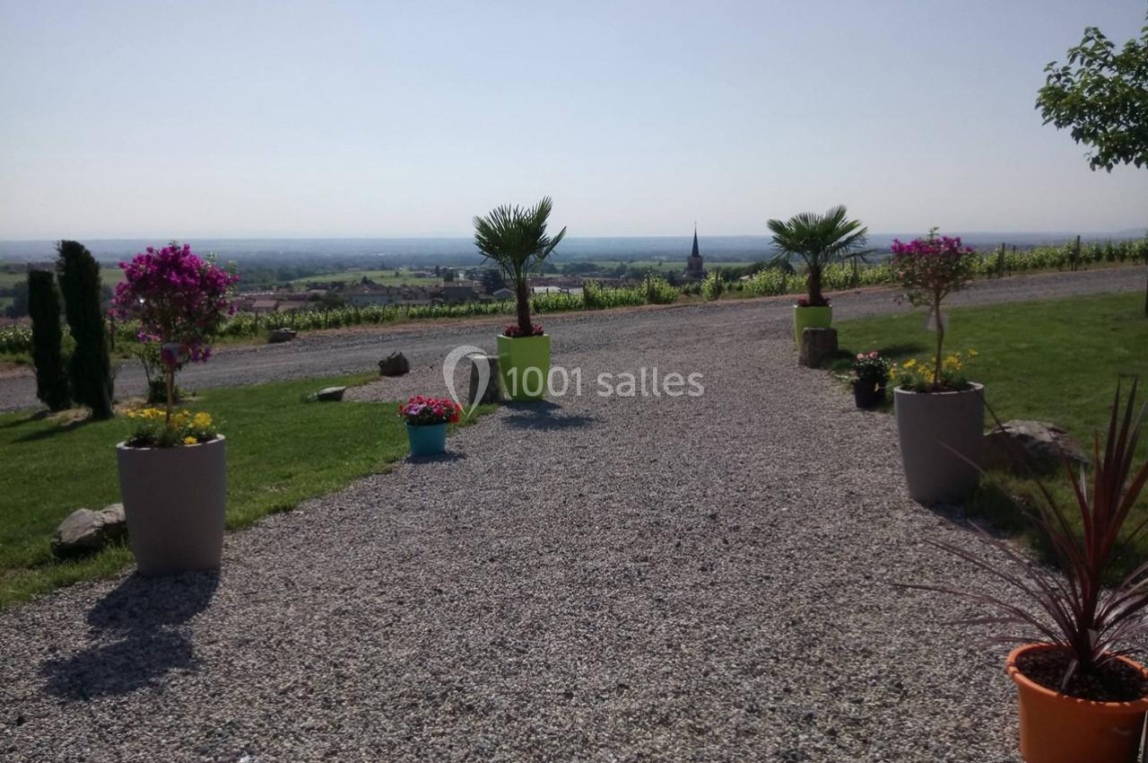 Allée en gravier bordée de pots de fleurs colorés, menant à un paysage de vignobles et une église au loin.