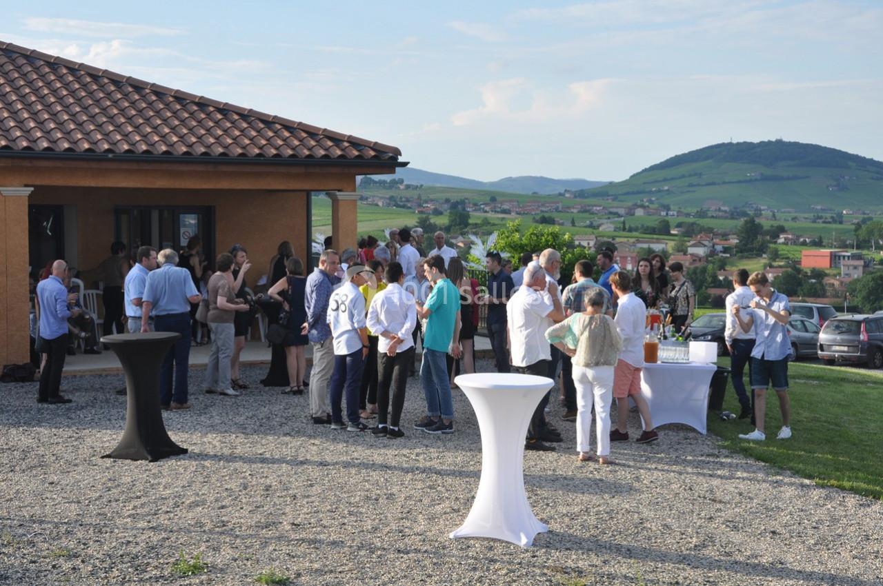 Groupe de personnes rassemblées en extérieur près d'une maison, discutant autour de tables hautes, avec un paysage vallonné…