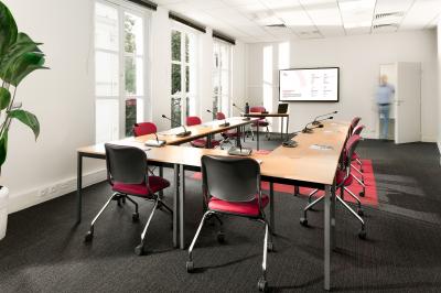 Salle de réunion lumineuse avec tables en U, chaises rouges, micros, écran mural et une plante verte.