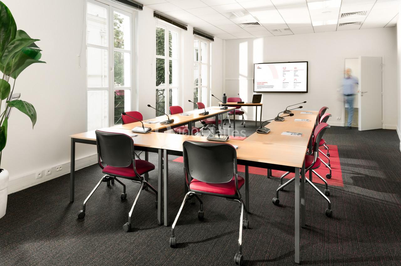 Salle de réunion lumineuse avec tables en U, chaises rouges, micros, écran mural et une plante verte.