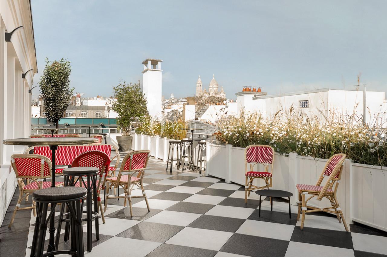 Terrasse avec sol en damier, tables et chaises rouges, vue sur des toits et la basilique du Sacré-Cœur au loin.