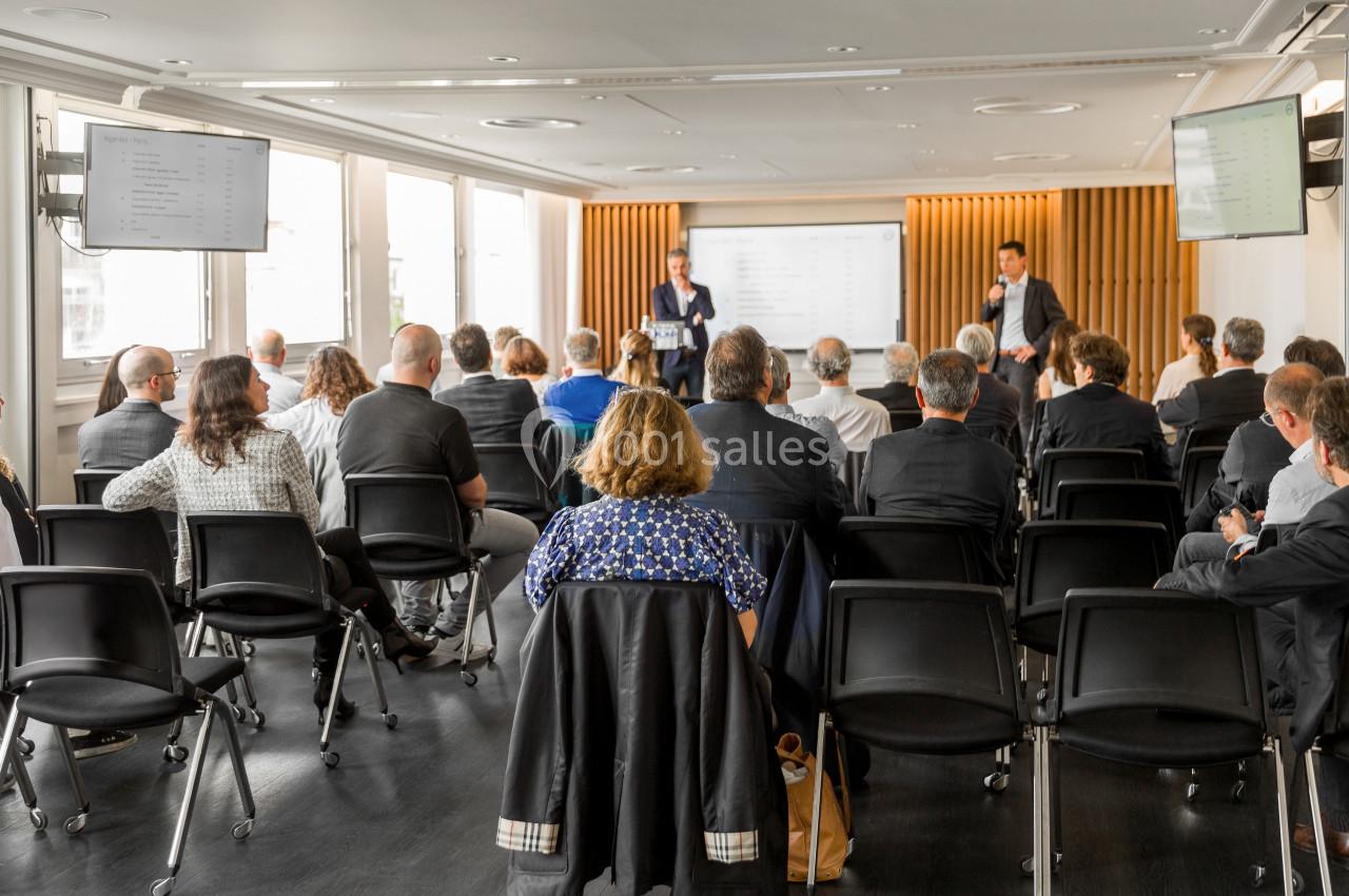 Un groupe de personnes assises dans une salle de conférence, écoutant deux intervenants devant un écran.