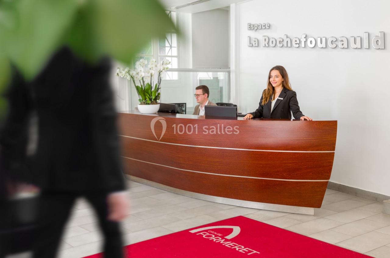 Accueil d'un espace professionnel avec un comptoir en bois, une réceptionniste souriante et un homme en arrière-plan.