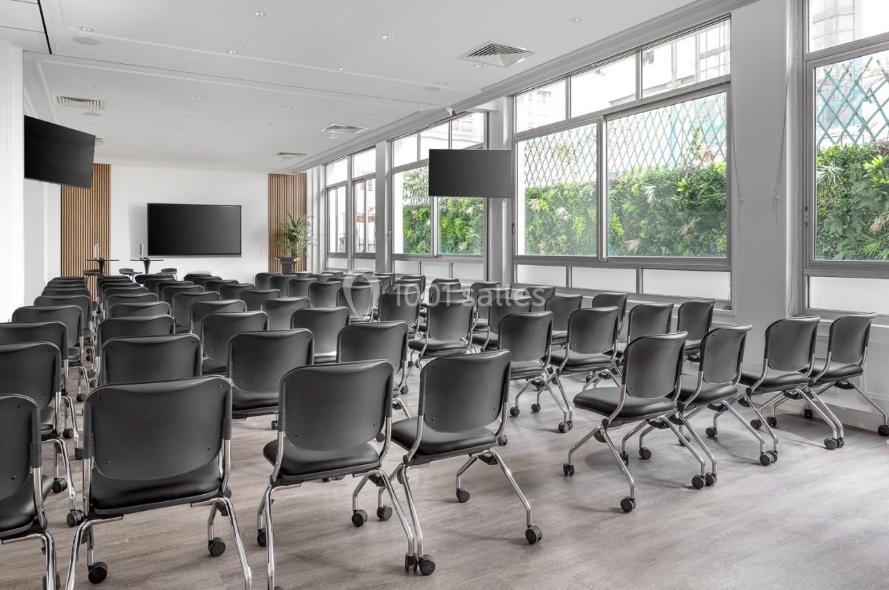 Salle de conférence lumineuse avec des rangées de chaises noires, écrans et grandes fenêtres donnant sur un espace vert.