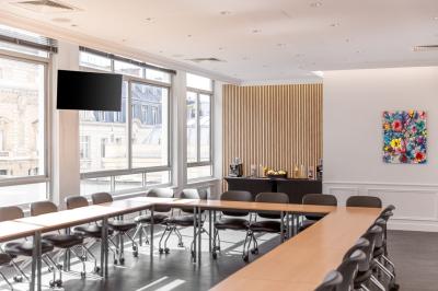 Salle de réunion lumineuse avec tables en U, chaises rouges, micros, écran mural et une plante verte.
