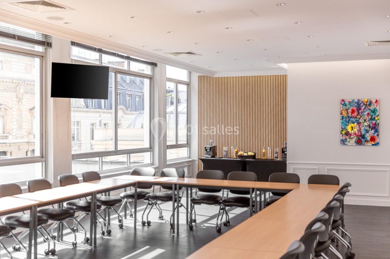 Salle de réunion lumineuse avec grandes fenêtres, tables en U, chaises noires et coin café en arrière-plan.