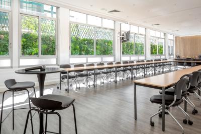 Salle de réunion lumineuse avec tables en U, chaises rouges, micros, écran mural et une plante verte.