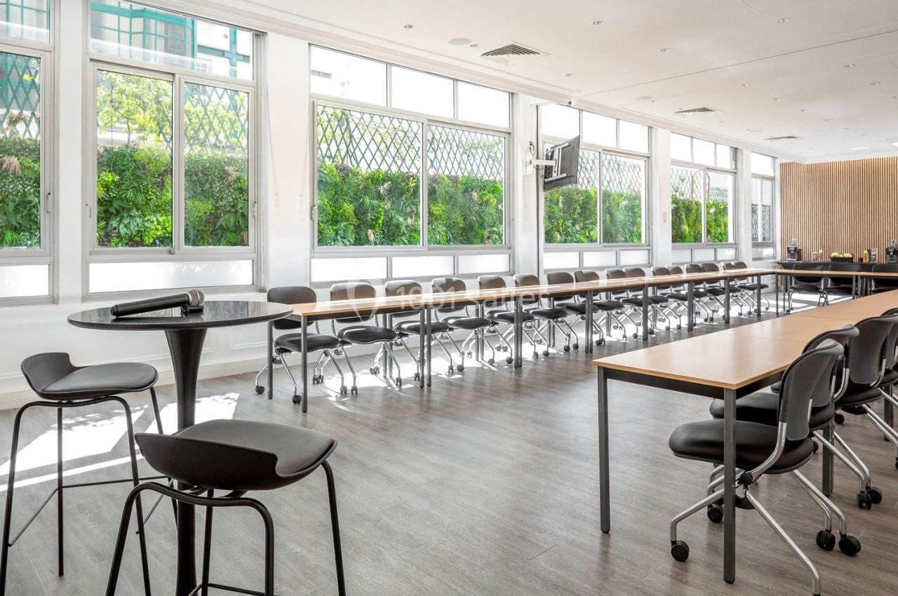 Salle de réunion lumineuse avec grandes fenêtres, tables en bois, chaises noires et sol en parquet gris.