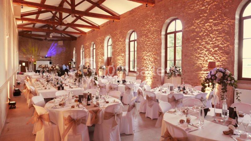 Salle de réception lumineuse avec murs en pierre, tables décorées de nappes blanches et compositions florales.