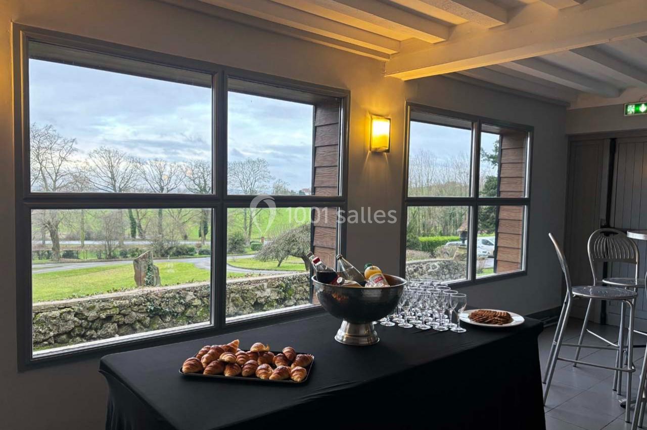 Table avec viennoiseries, fruits et verres, placée devant de grandes fenêtres offrant une vue sur un paysage verdoyant.