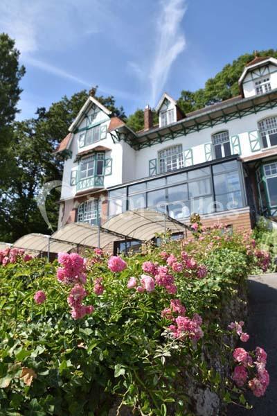 Grande maison blanche à colombages entourée de rosiers fleuris, avec ciel bleu et arbres en arrière-plan.