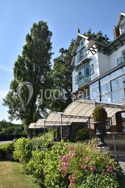 Façade d'une maison à colombages avec verrière, entourée de jardins fleuris et d'arbres sous un ciel bleu.