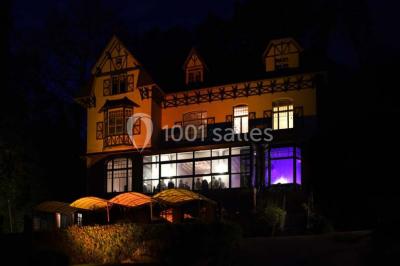 Grande maison à colombages éclairée en vert la nuit, avec des fenêtres illuminées et un porche couvert.