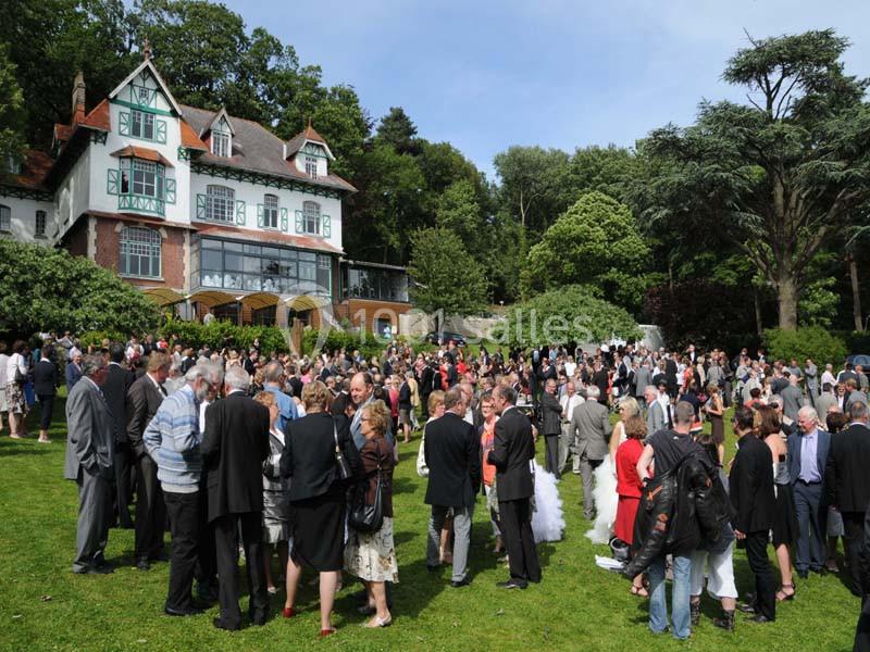 Groupe de personnes rassemblées dans un jardin devant une grande maison, lors d'un événement en plein air.