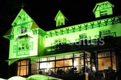 Grande maison à colombages éclairée en vert la nuit, avec des fenêtres illuminées et un porche couvert.