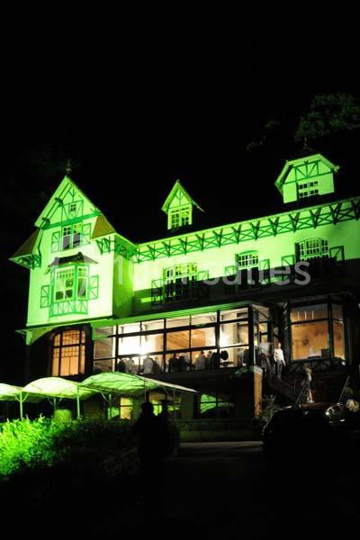 Grande maison à colombages éclairée en vert la nuit, avec des fenêtres illuminées et un porche couvert.