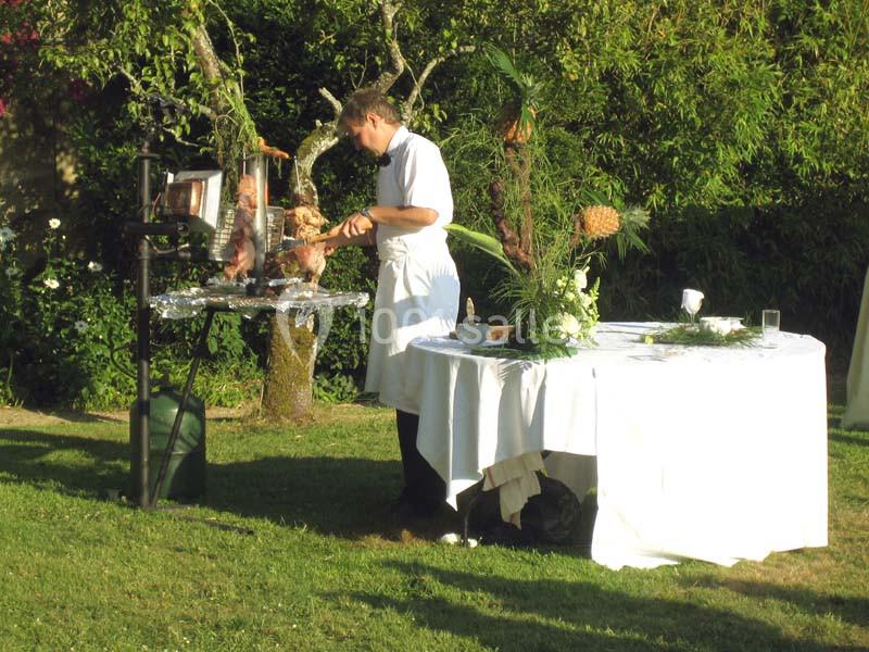 Un cuisinier en tablier prépare un rôti sur une broche dans un jardin, près d'une table décorée de fleurs.