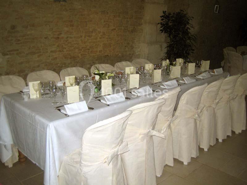 Table dressée avec nappes blanches, chaises couvertes, menus, verres et couverts dans une salle aux murs en pierre.