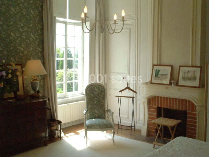 Chambre lumineuse avec fauteuil, cheminée, commode, et grande fenêtre donnant sur un extérieur verdoyant.